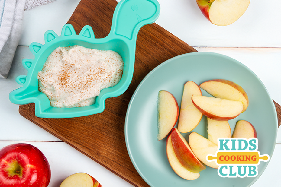 apple slices on a plate with a yogurt dip in a dinosaur-shaped bowl on a wooden cutting board
