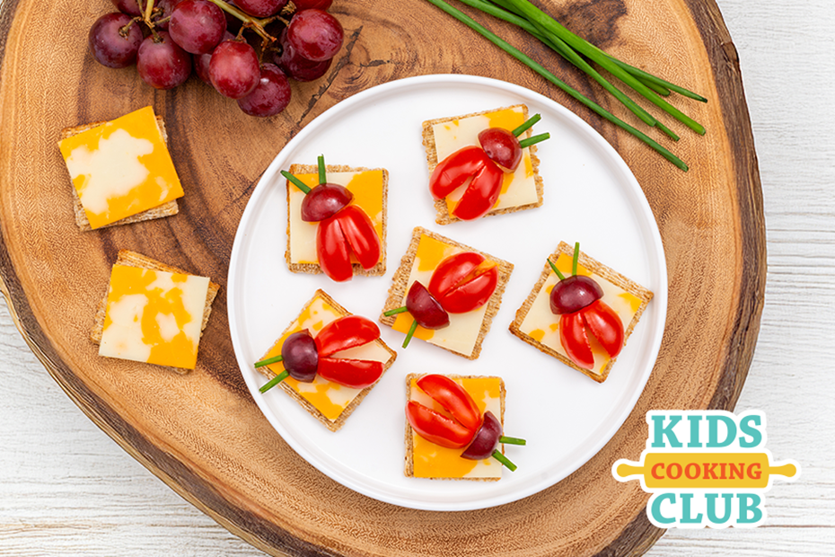 ladybugs made out of a cracker, cheese, tomatoes as the body and a grape for the head on a white plate. This is on a wooden cutting board with a bunch of grapes and some cheese beside it.  