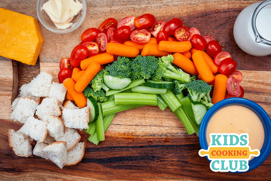 rainbow made with pieces of bread and fresh veggies with a bowl of cheese dip at the end on a wooden board. Milk and cheese beside it.  